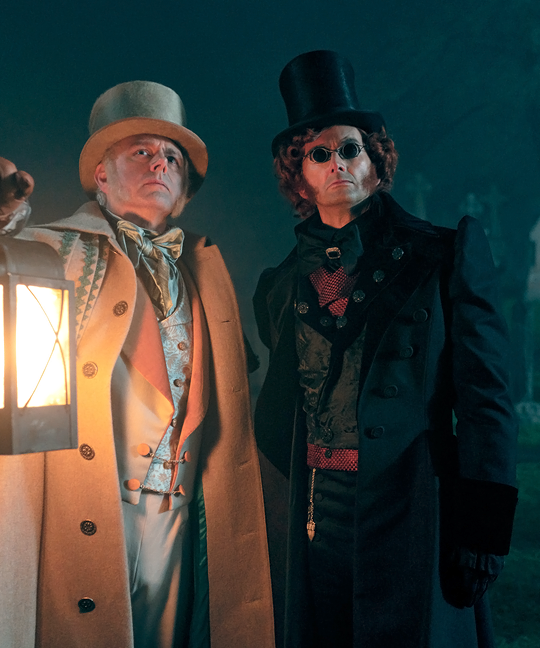 Aziraphale and Crowley in a graveyard in regency Edinburgh. Aziraphale holds aloft a lantern.