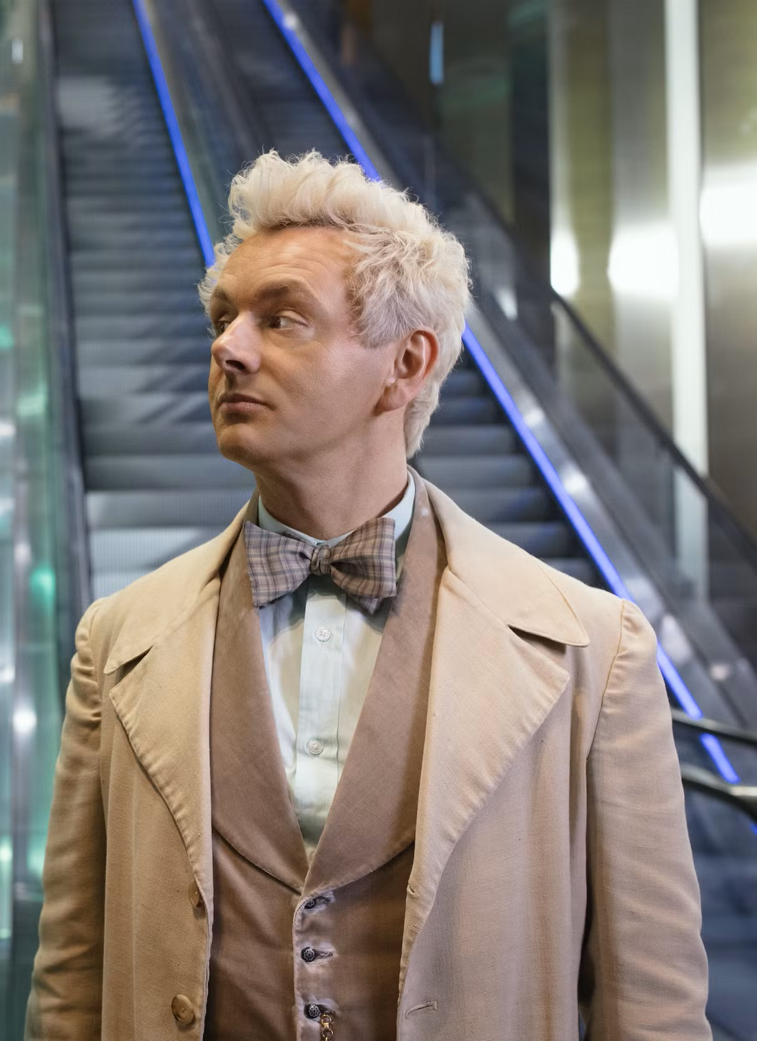 Aziraphale standing at the bottom of an escalator looking to his right.