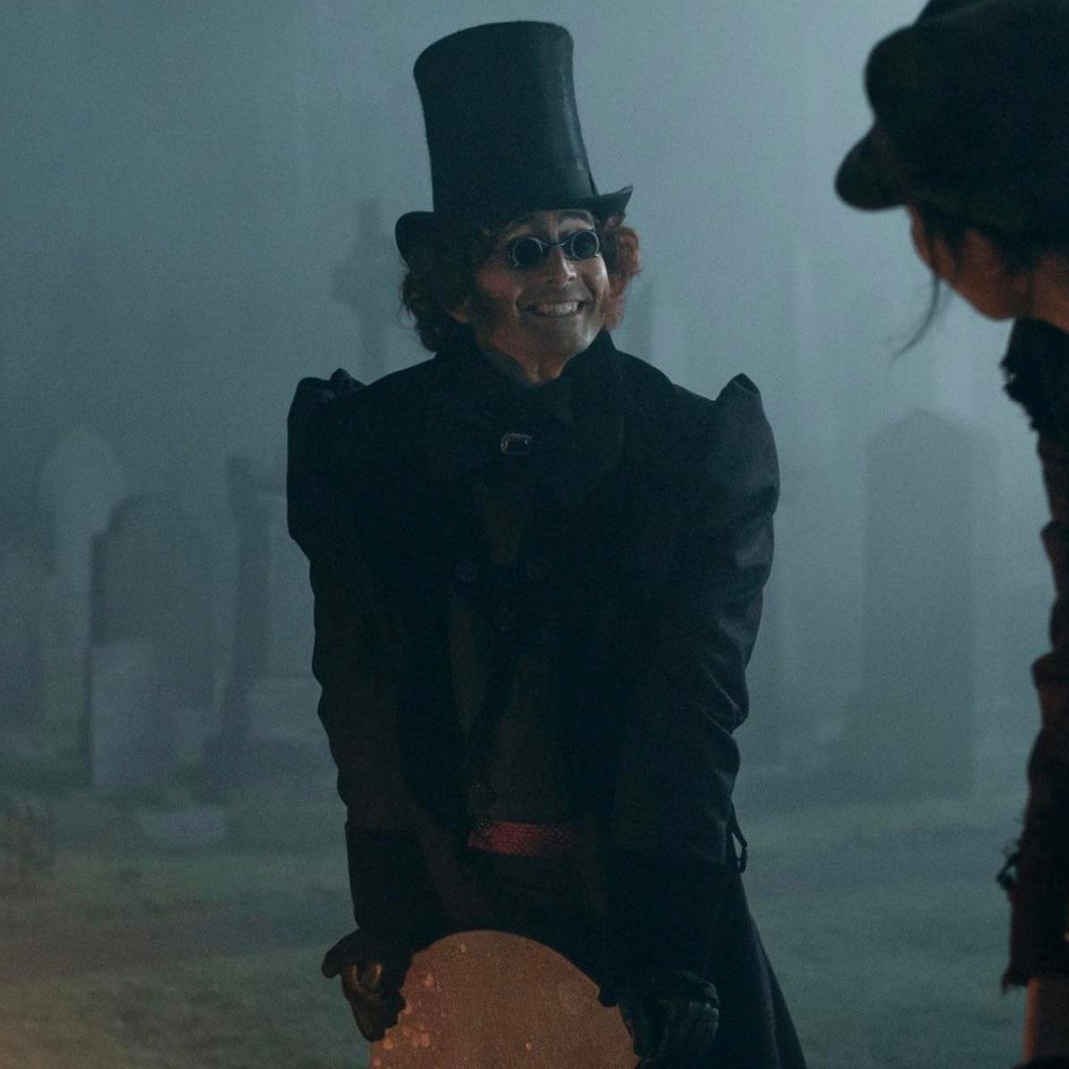 Crowley, in his regency attire, leans against a gravestone with a large smile on his face.