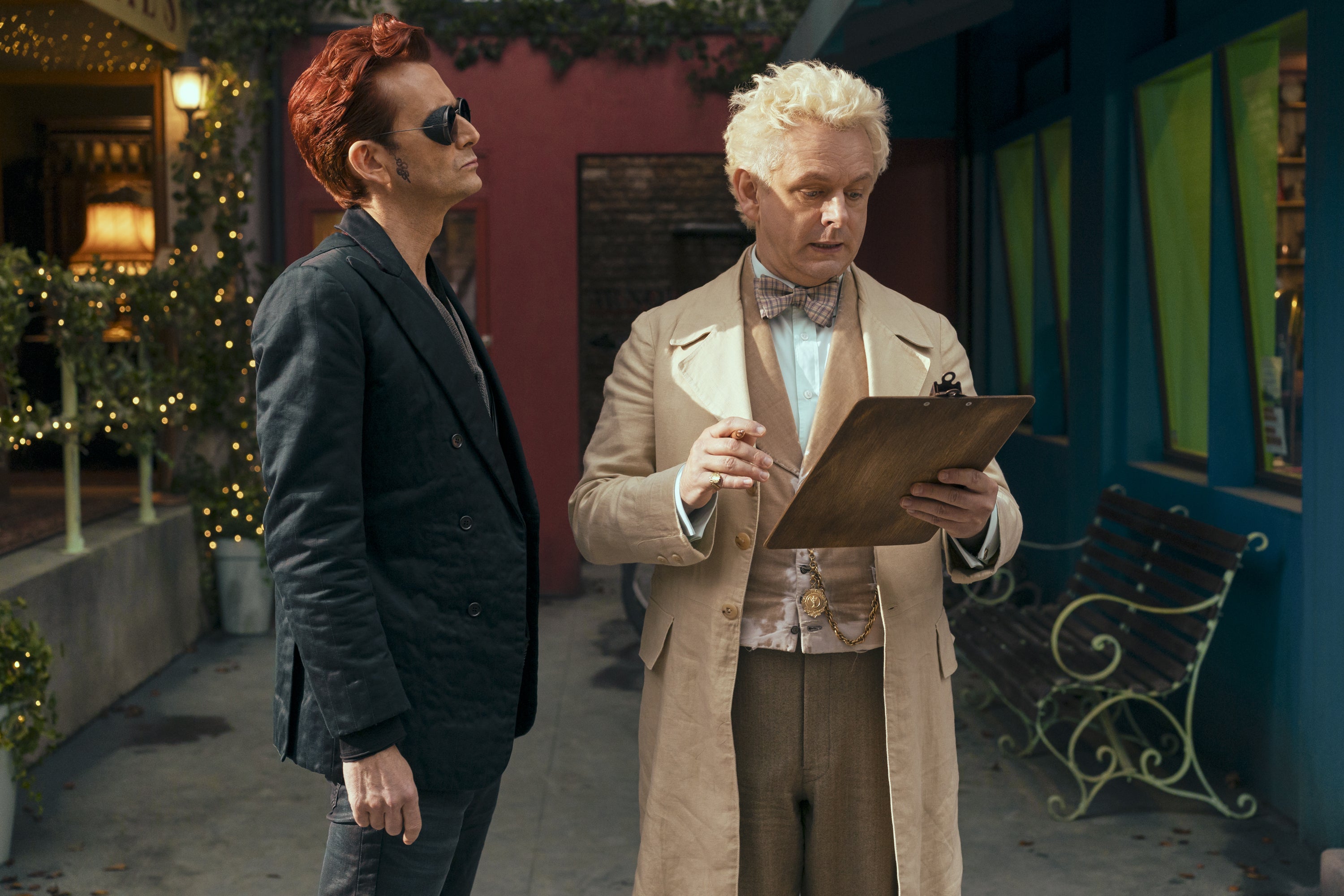 Crowley standing next to Aziraphale as he looks at his clipboard.