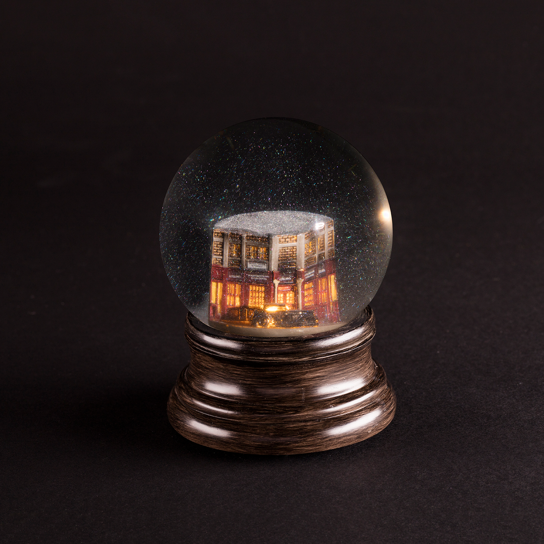 A snowglobe featuring Aziraphale's bookshop and Crowley's Bentley. The bookshop is ablaze, leading to a smoke effect. Photographed against a black backdrop.