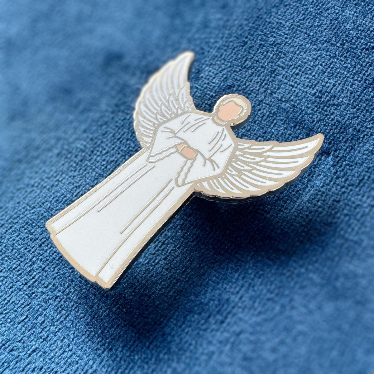 Photograph of a Good Omens enamel pin on a fabric backdrop: Angelic Aziraphale