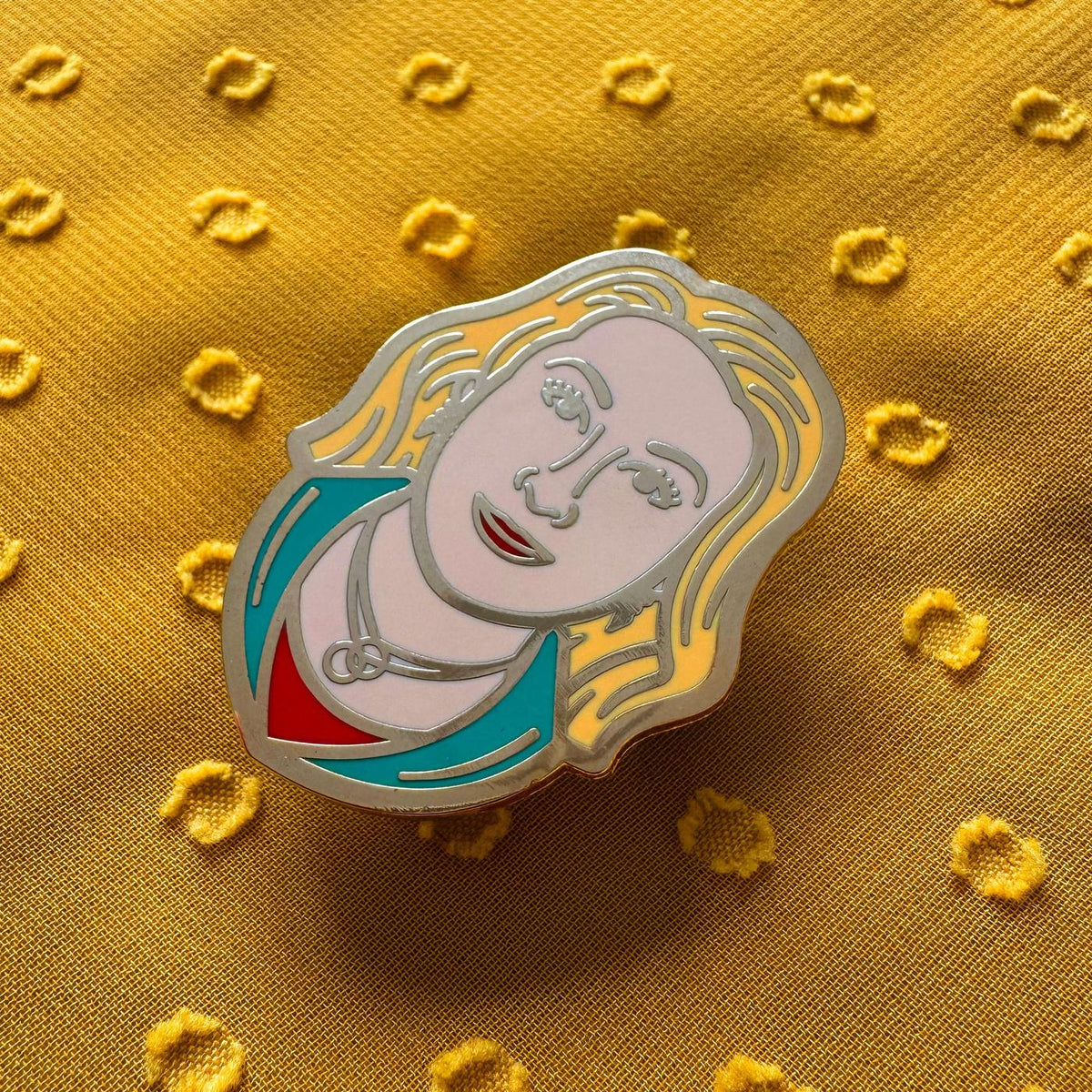 Photograph of a Good Omens enamel pin on a fabric background: Maggie